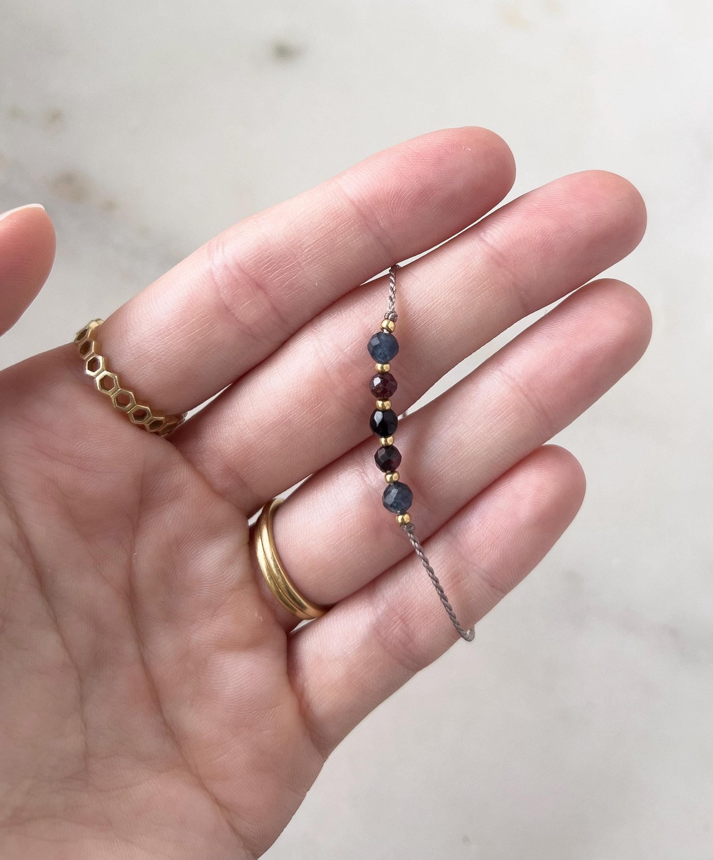 Dlaň ženské ruky, která drží mezi prsty minimalistický náramek vytvořený z šedé hedvábné nitě, navlečený pěti minerály (modrý safír, červený granát, černý onyx, a další granát a safír), mezi kterými jsou další malé zlaté korálky.