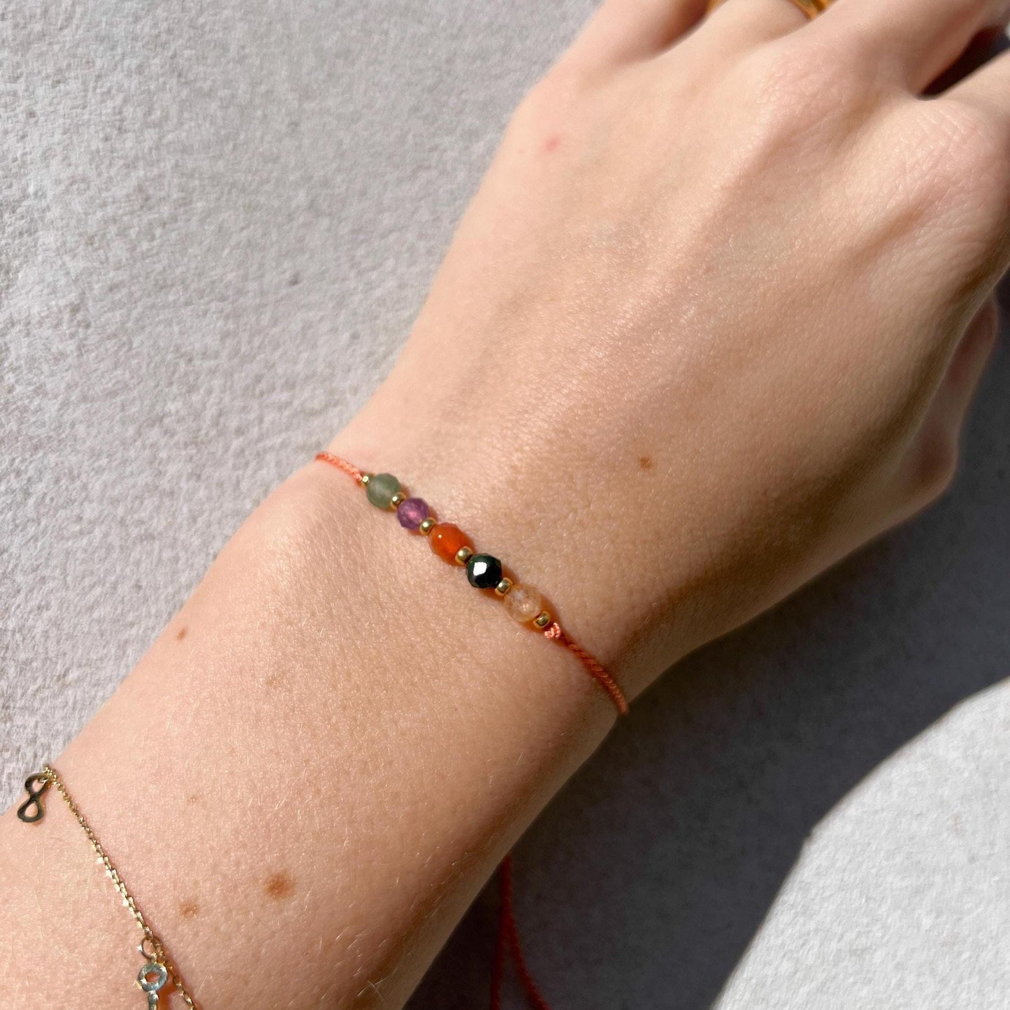 Dívčí ruka, která má na sobě tenký oranžový hedvábný náramek s těmito minerály: žlutý citrín, zelený jadeit, oranžový karneol, fialový ametyst, zelený avanturín. Mezi minerály jsou umístěny malé zlaté korálky, které dodávají náramku eleganci.