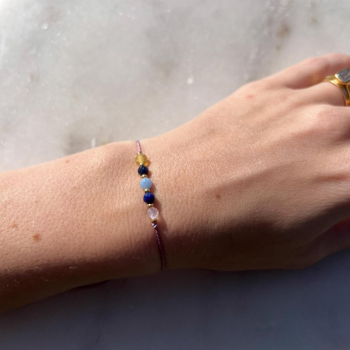 Dívčí ruka, která má na sobě tenký fialový hedvábný náramek s těmito minerály: žlutý opál, modrý lapis lazuli, modrý akvamarín, další lapis lazuli a růženín. Mezi minerály jsou umístěny malé zlaté korálky, které dodávají náramku eleganci.