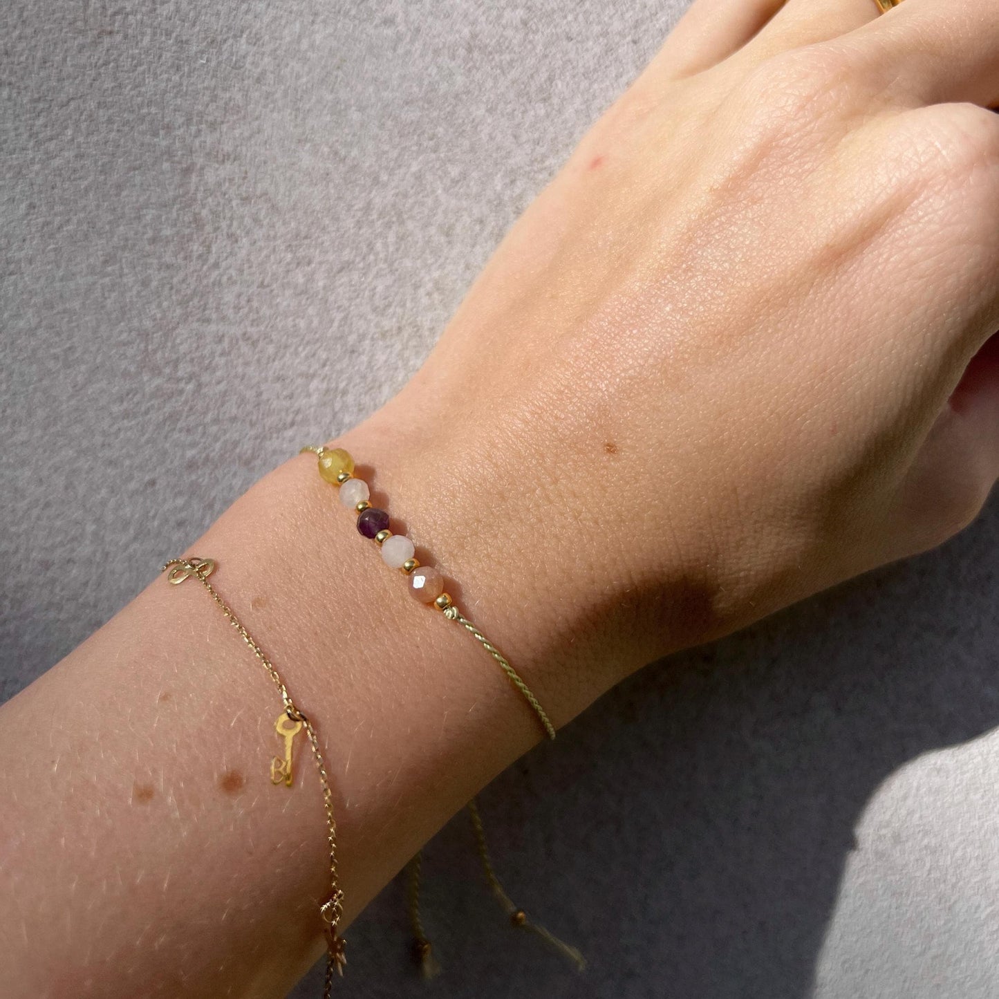 Dívčí ruka, která má na sobě tenký zelený hedvábný náramek s těmito minerály: ametyst, měsíční kámen, opál, sluneční kámen, růženín. Mezi minerály jsou umístěny malé zlaté korálky, které dodávají náramku eleganci. Na ruce je také další jemný zlatý náramek, se kterým se minerální náramek hezky doplňuje.