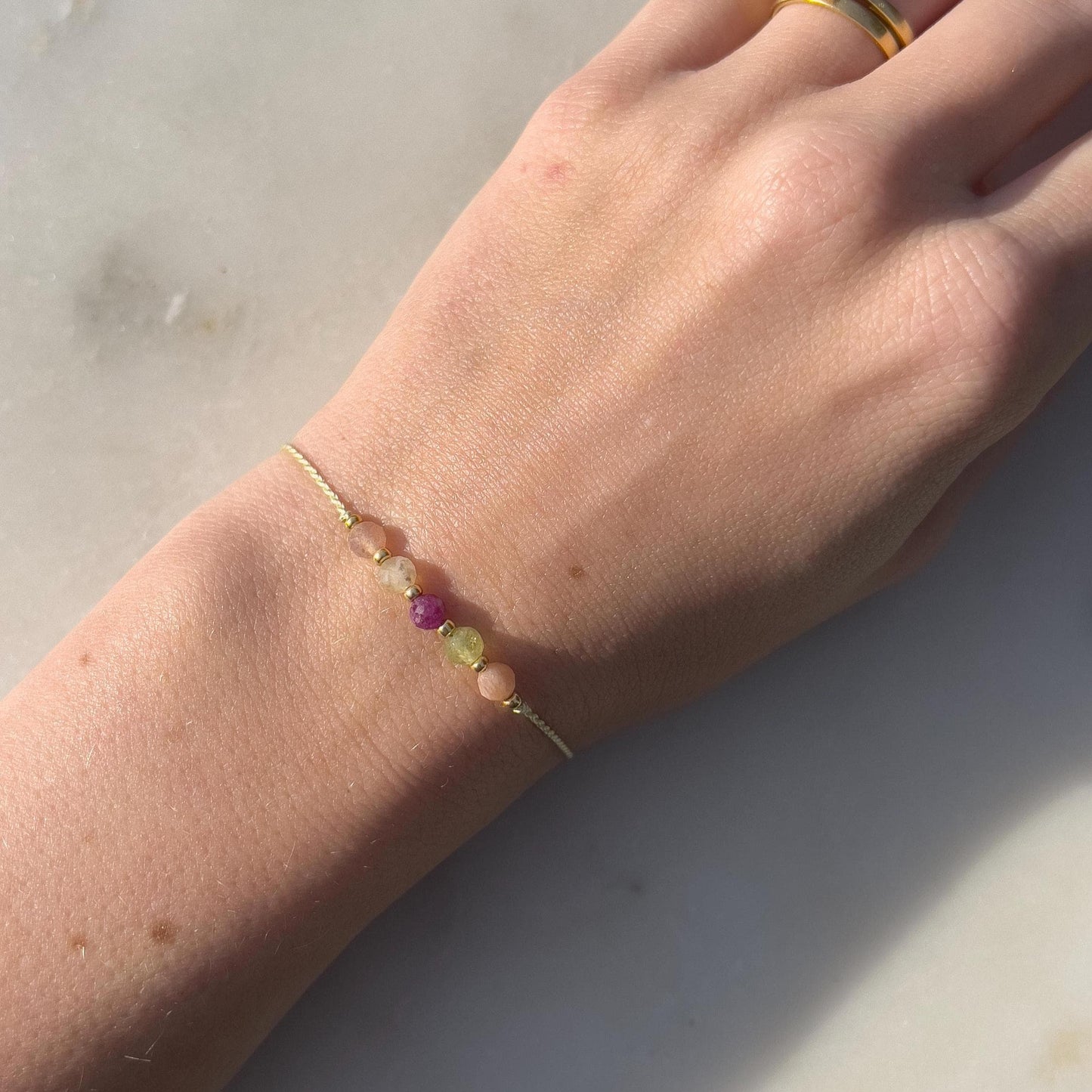 Dívčí ruka, která má na sobě tenký zelený hedvábný náramek s těmito minerály: sluneční kámen, zelený olivín, červený rubín, žlutý citrín a další sluneční kámen. Mezi minerály jsou umístěny malé zlaté korálky, které dodávají náramku eleganci.