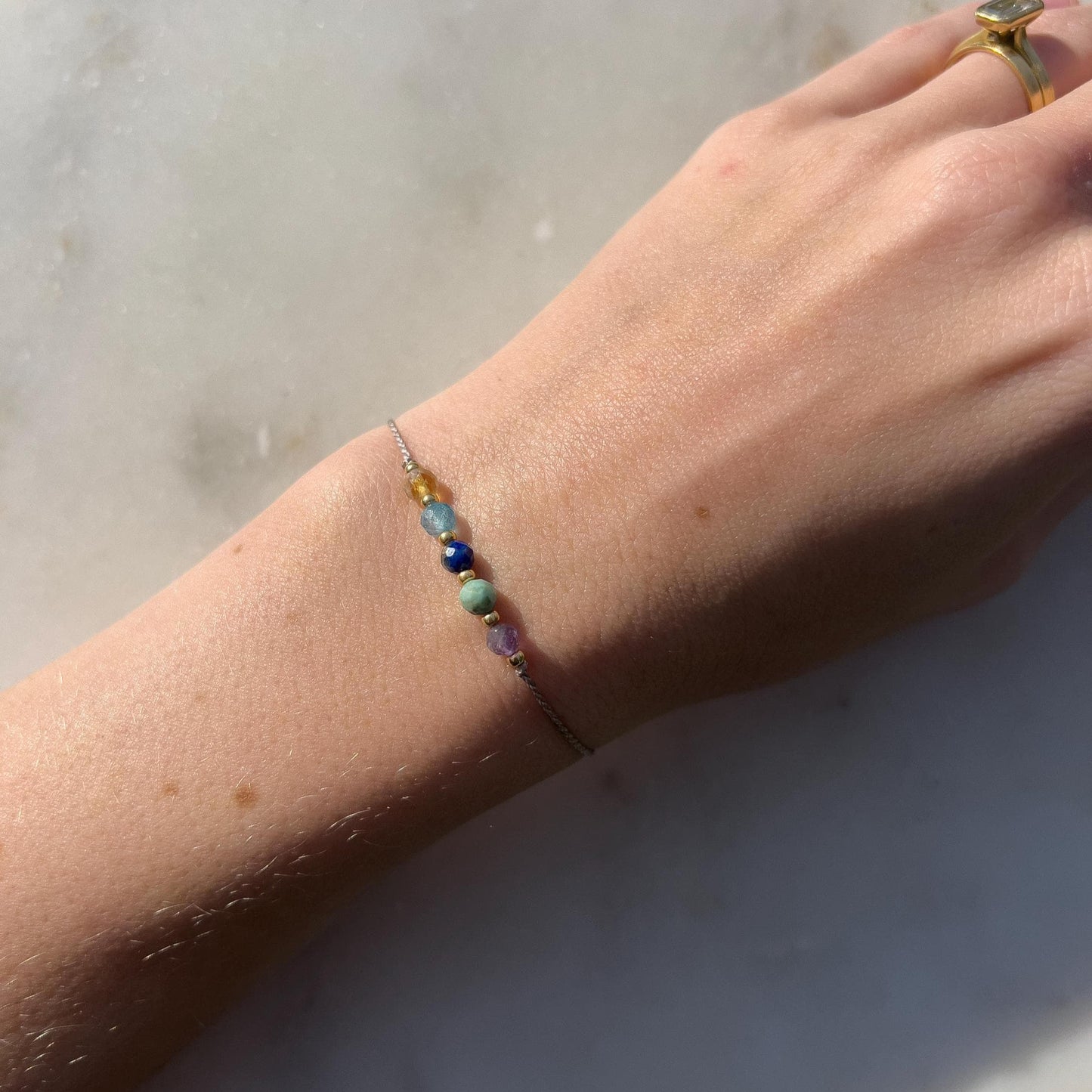 Dívčí ruka, která má na sobě tenký šedý hedvábný náramek s těmito minerály: fialový ametyst, zelený tyrkys, modrý lapis lazuli, modrý topaz a žlutý citrín. Mezi minerály jsou umístěny malé zlaté korálky, které dodávají náramku eleganci.