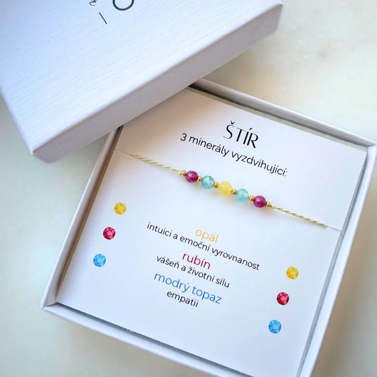 Otevřená bílá krabička s jemným náramkem pro znamení Štír. Náramek na zelené šňůrce zdobí pět korálků z přírodních minerálů (dva rubíny, dva modré topazy a opál). Význam těchto minerálů je napsán na kartičce, která je součástí krabičky.