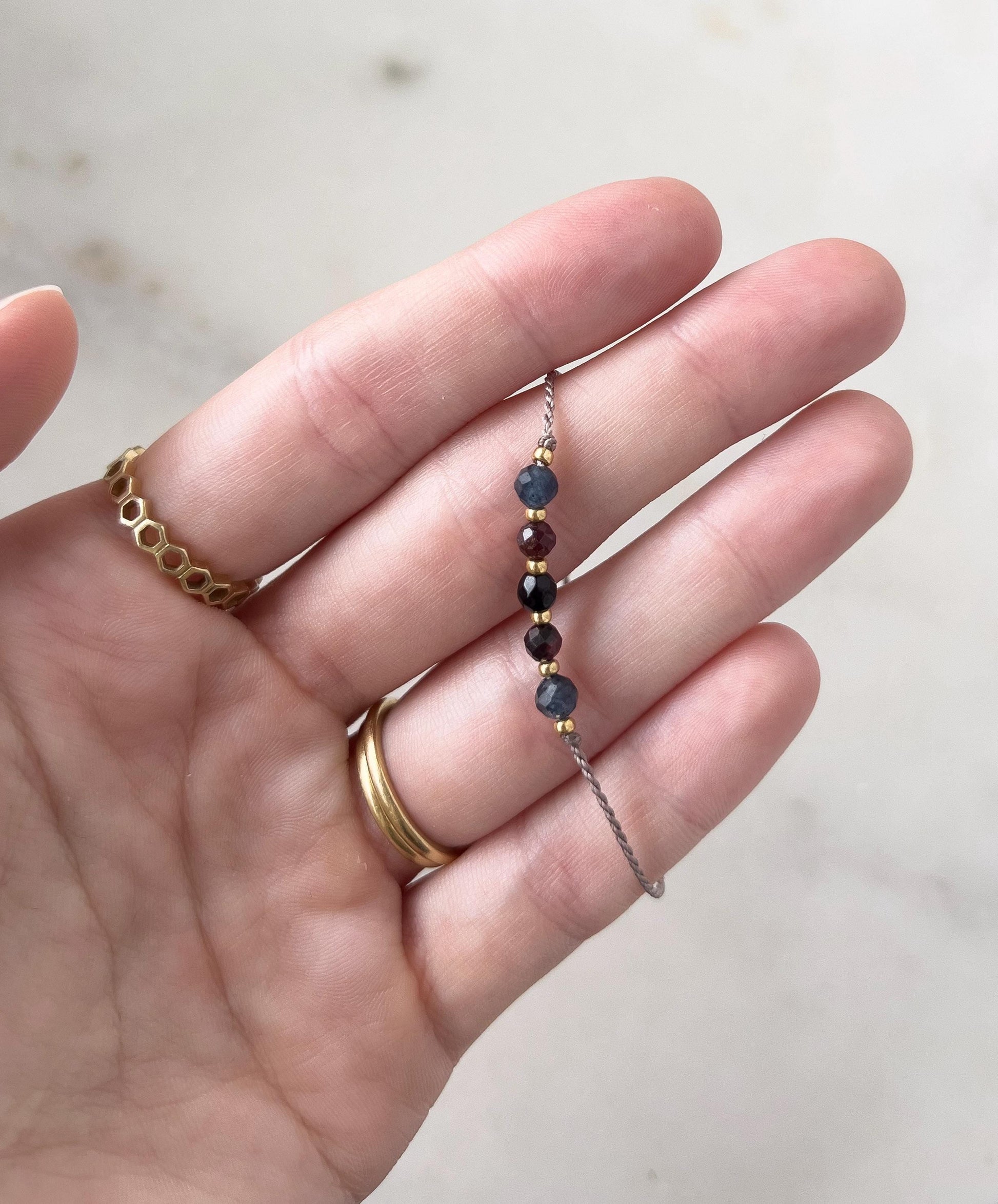 Dlaň ženské ruky, která drží mezi prsty minimalistický náramek vytvořený z šedé hedvábné nitě, navlečený pěti minerály (modrý safír, červený granát, černý onyx, a další granát a safír), mezi kterými jsou další malé zlaté korálky.