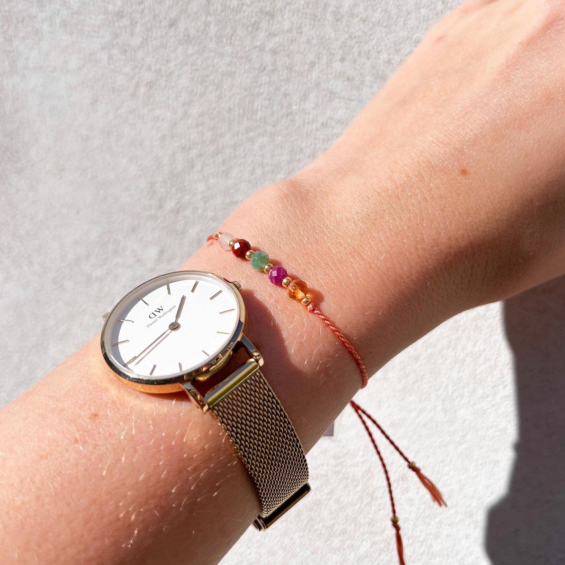 Dívčí ruka, která má na sobě tenký oranžový hedvábný náramek s těmito minerály: měsíční kámen, červený jaspis, zelený smaragd, růžový rubín, žlutý citrín. Mezi minerály jsou umístěny malé zlaté korálky, které dodávají náramku eleganci. Modelka má na ruce také klasické DW kulaté hodinky, se zlatým kovovým páskem, čímž ukazuje jak náramek hezky doplňuje a ladí s hodinkami.