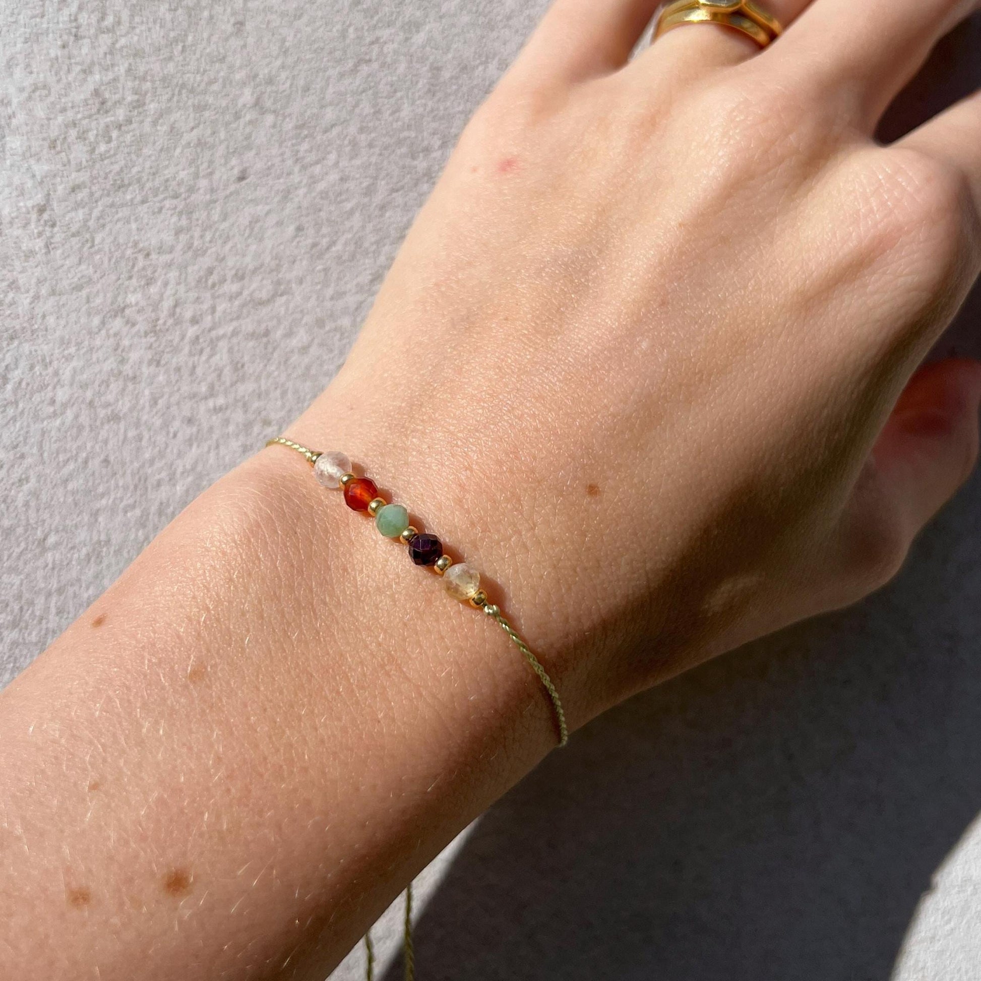 Dívčí ruka, která má na sobě tenký zelený hedvábný náramek s těmito minerály: křišťál, oranžový karneol, zelený smaragd, červený granát, žlutý citrín. Mezi minerály jsou umístěny malé zlaté korálky, které dodávají náramku eleganci.