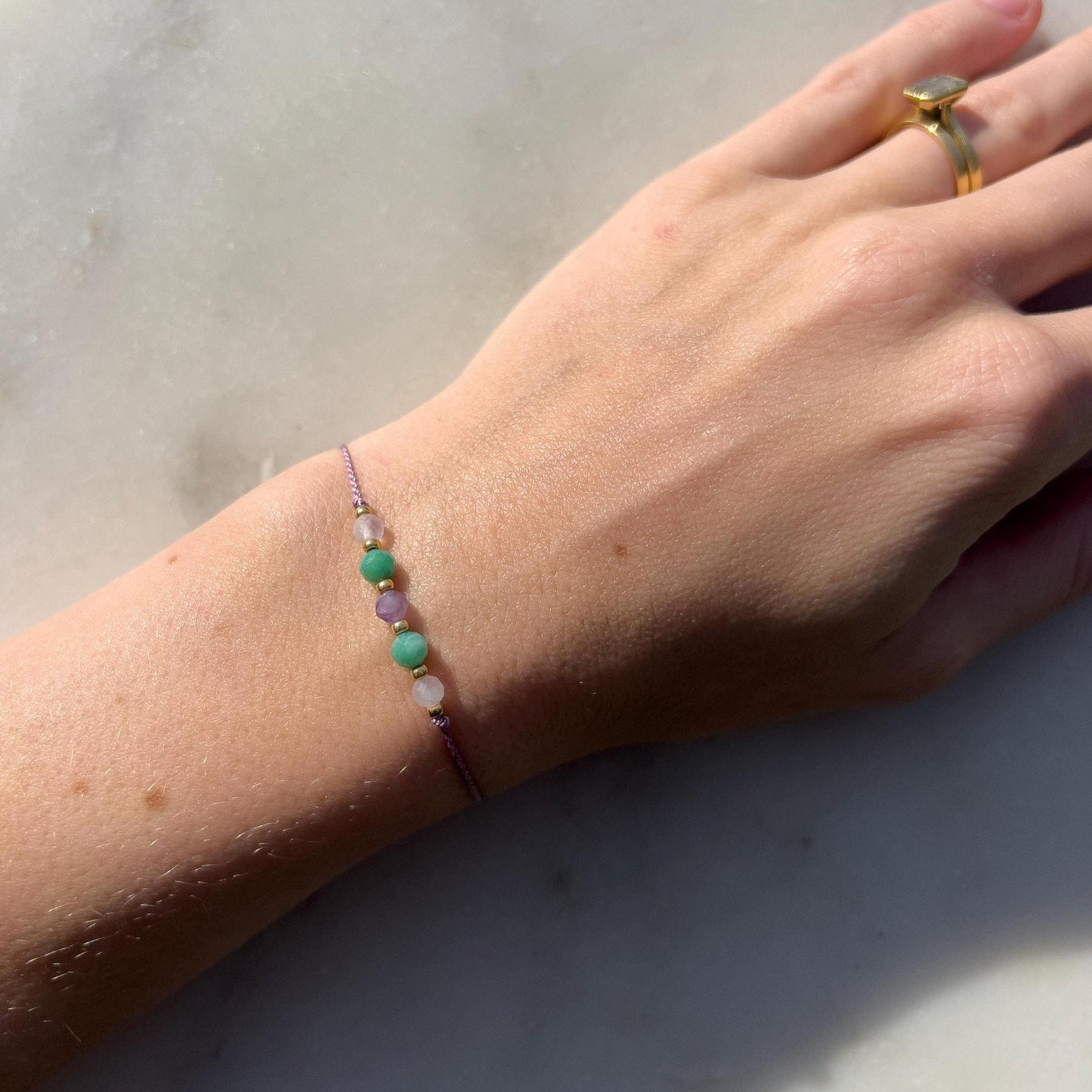 Dívčí ruka, která má na sobě tenký oranžový hedvábný náramek s těmito minerály: měsíční kámen, modrý amazonit, fialový ametyst, další amazonit a měsíční kámen. Mezi minerály jsou umístěny malé zlaté korálky, které dodávají náramku eleganci.