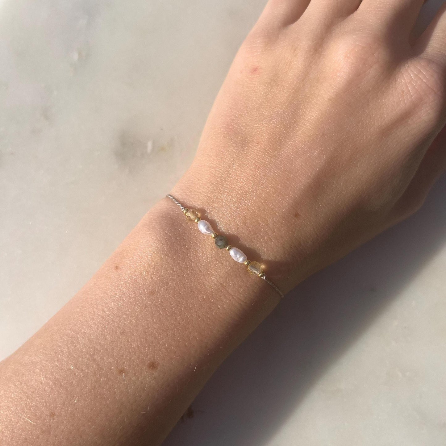 Dívčí ruka, která má na sobě tenký šedý hedvábný náramek s těmito minerály: žlutý citrín, bílá perla, zelený smaragd, další perla a citrín. Mezi minerály jsou umístěny malé zlaté korálky, které dodávají náramku eleganci.