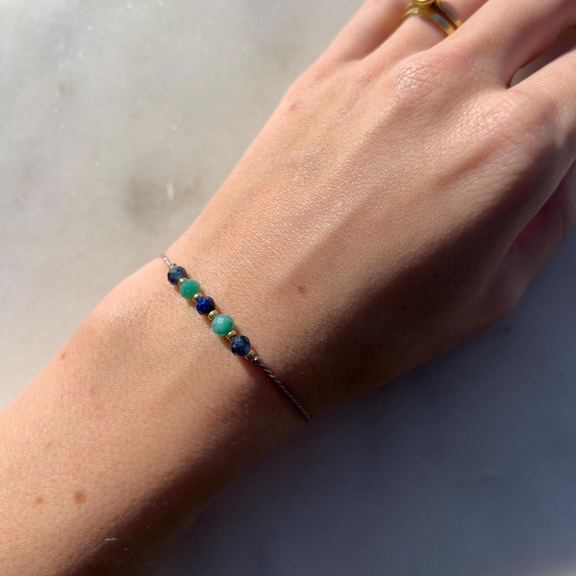 Dívčí ruka, která má na sobě tenký šedý hedvábný náramek s těmito minerály: safír, amazonit, lapis lazuli, další amazonit a safír. Mezi minerály jsou umístěny malé zlaté korálky, které dodávají náramku eleganci.