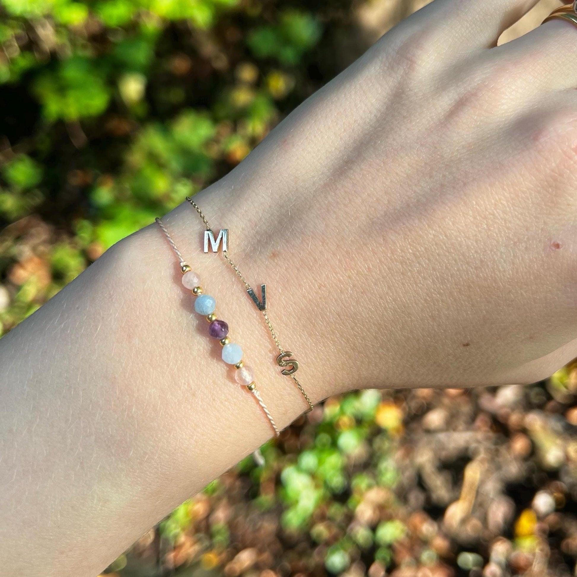 Ženská ruka, která má na sobě růžový hedvábný náramek s těmito minerály: růženín, modrý akvamarín, fialový ametyst, další akvamarín a růženín. Mezi minerály jsou umístěny malé zlaté korálky, které dodávají náramku eleganci. Společně s tímto náramkem je na ruce druhý jemný zlatý náramek se třemi iniciály MVS, což ukazuje jak lze minerální náramek kombinovat s dalšími šperky.