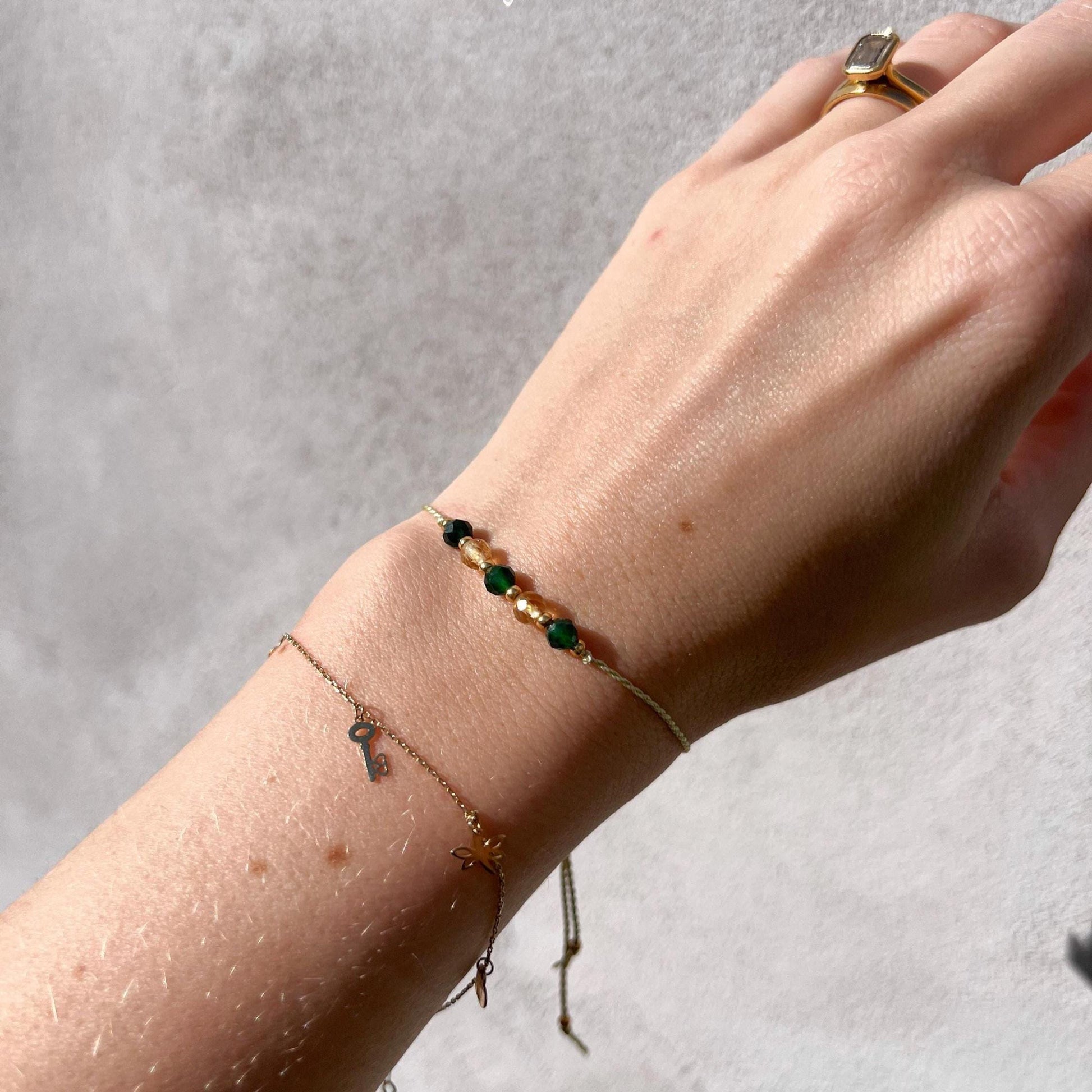 Dívčí ruka, která má na sobě tenký zelený hedvábný náramek s těmito minerály: střídavě tři zelené jadeity, dva žluté citríny. Mezi minerály jsou umístěny malé zlaté korálky, které dodávají náramku eleganci. Na ruce je také další jemný zlatý náramek, který minerální náramek hezky doplňuje.