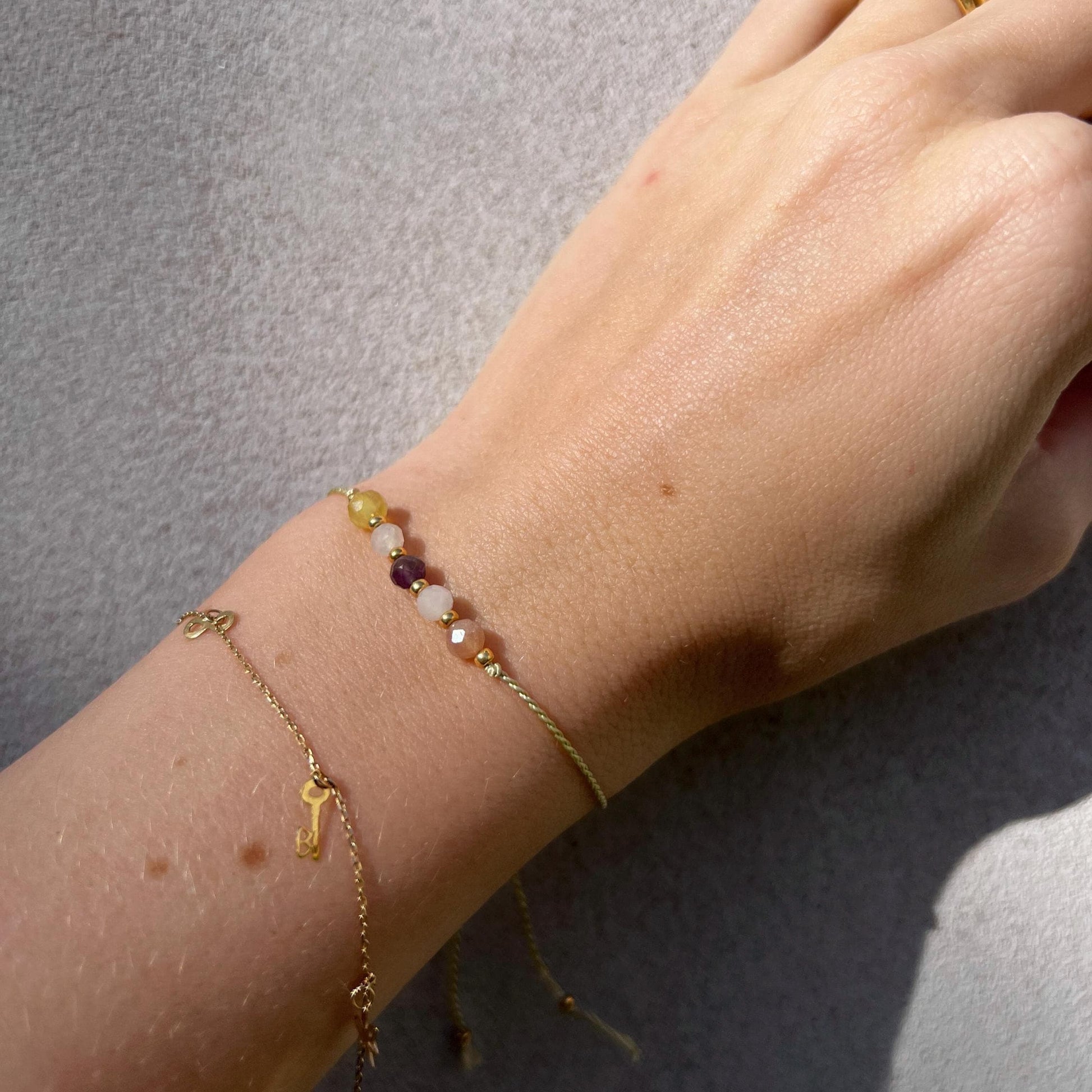 Dívčí ruka, která má na sobě tenký zelený hedvábný náramek s těmito minerály: ametyst, měsíční kámen, opál, sluneční kámen, růženín. Mezi minerály jsou umístěny malé zlaté korálky, které dodávají náramku eleganci. Na ruce je také další jemný zlatý náramek, se kterým se minerální náramek hezky doplňuje.