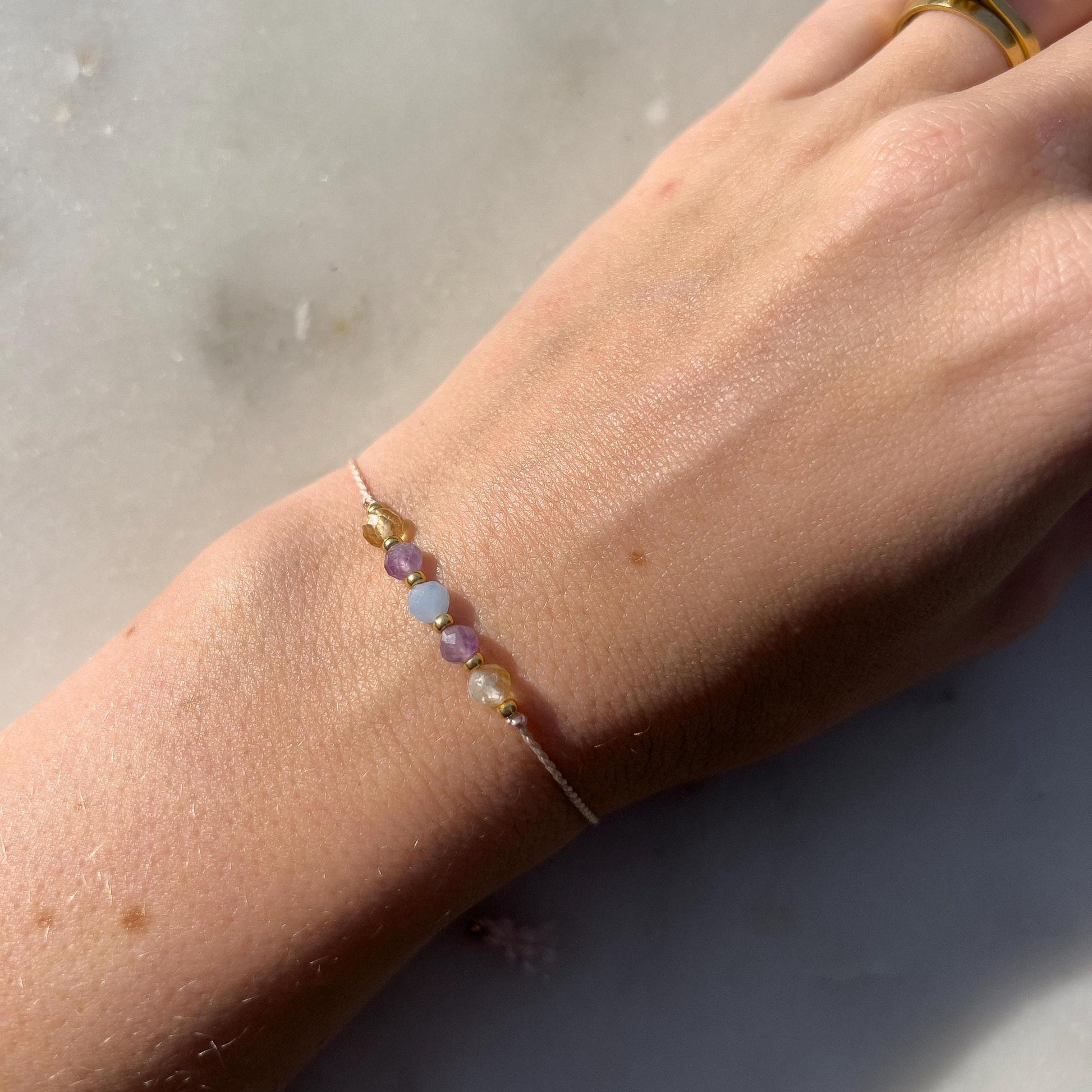 Dívčí ruka, která má na sobě tenký růžový hedvábný náramek s těmito minerály: žlutý citrín, fialový ametyst, modrý akvamarín, další ametyst a citrín. Mezi minerály jsou umístěny malé zlaté korálky, které dodávají náramku eleganci.