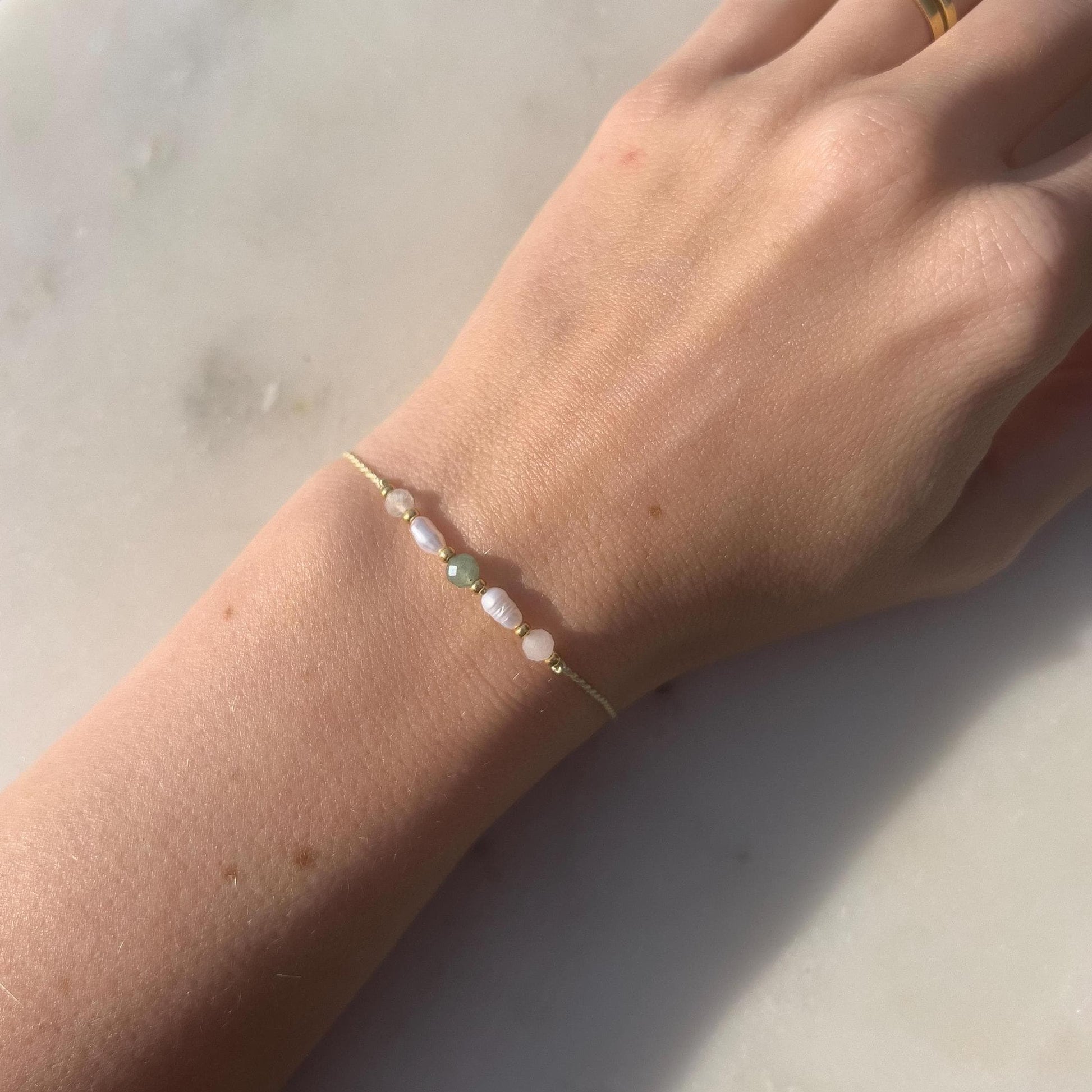 Dívčí ruka, která má na sobě tenký zelený hedvábný náramek s těmito minerály: měsíční kámen, bílá perla, zelený avanturín, další perla a růženín. Mezi minerály jsou umístěny malé zlaté korálky, které dodávají náramku eleganci.