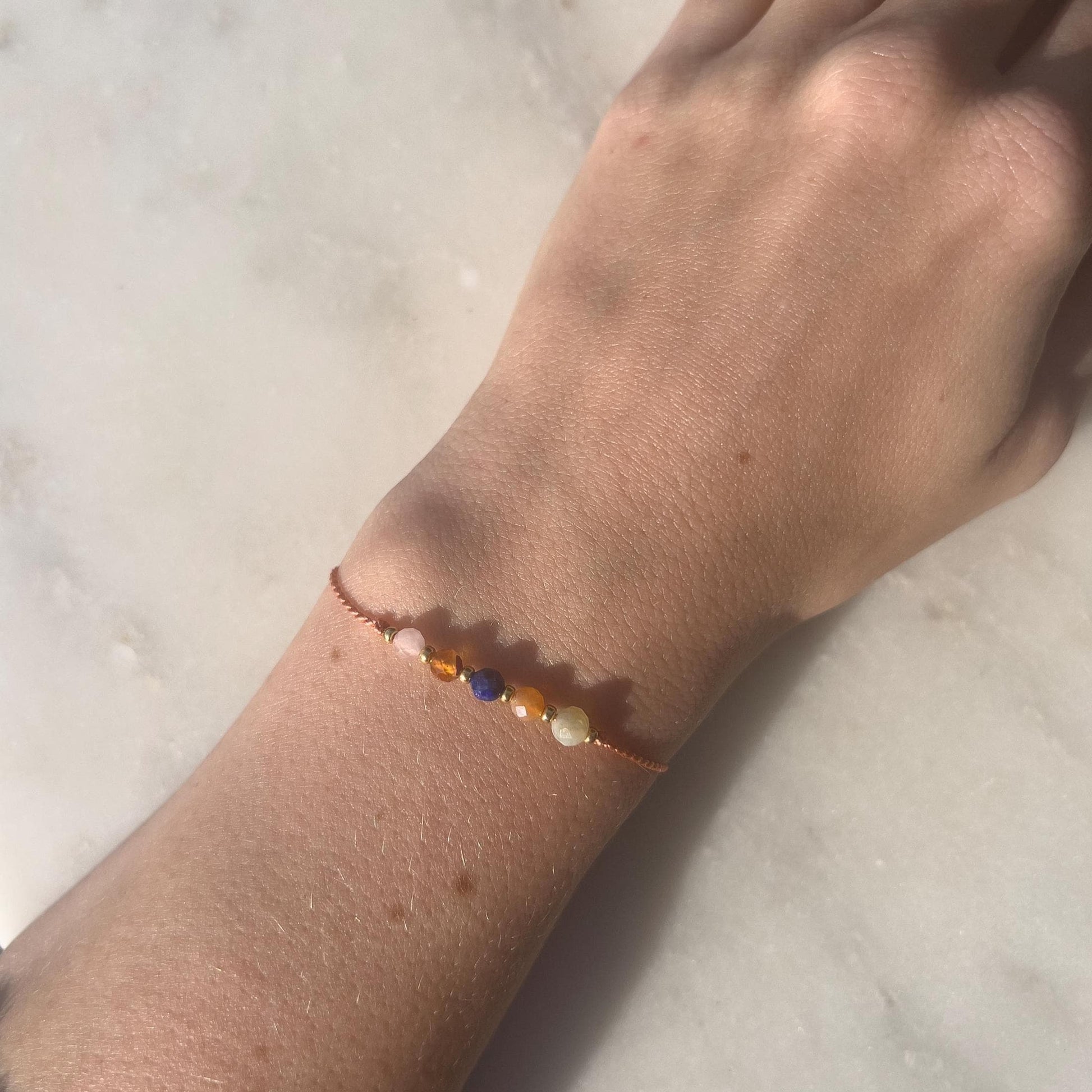 Dívčí ruka, která má na sobě tenký oranžový hedvábný náramek s těmito minerály: žlutý opál, oranžový karneol, modrý lapis lazuli, další karneol a růženín. Mezi minerály jsou také malé zlaté korálky, které dodávají náramku eleganci.