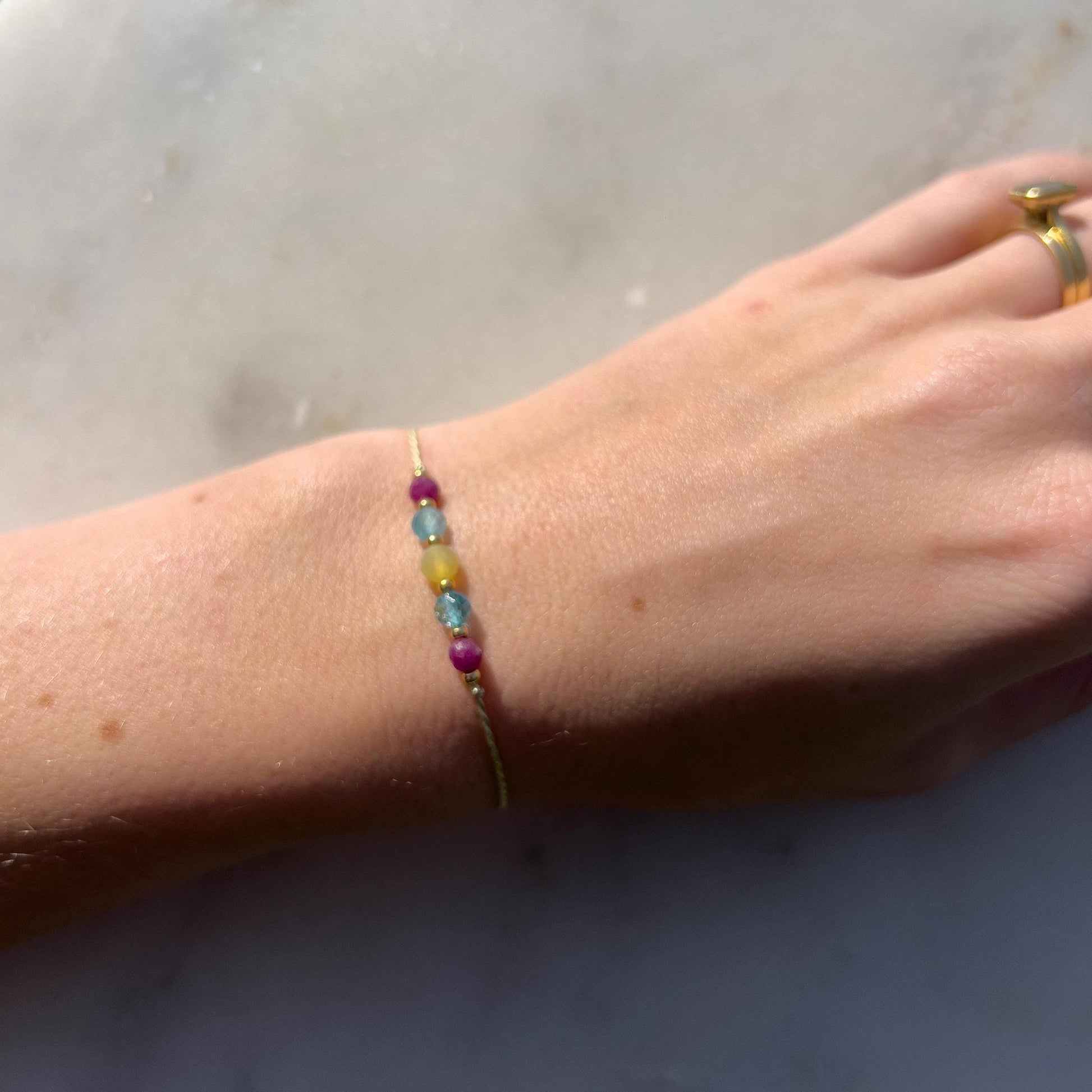 Zelený hedvábný náramek na ruce s pěti minerály: rubín, topaz, opál a zlaté korálky.